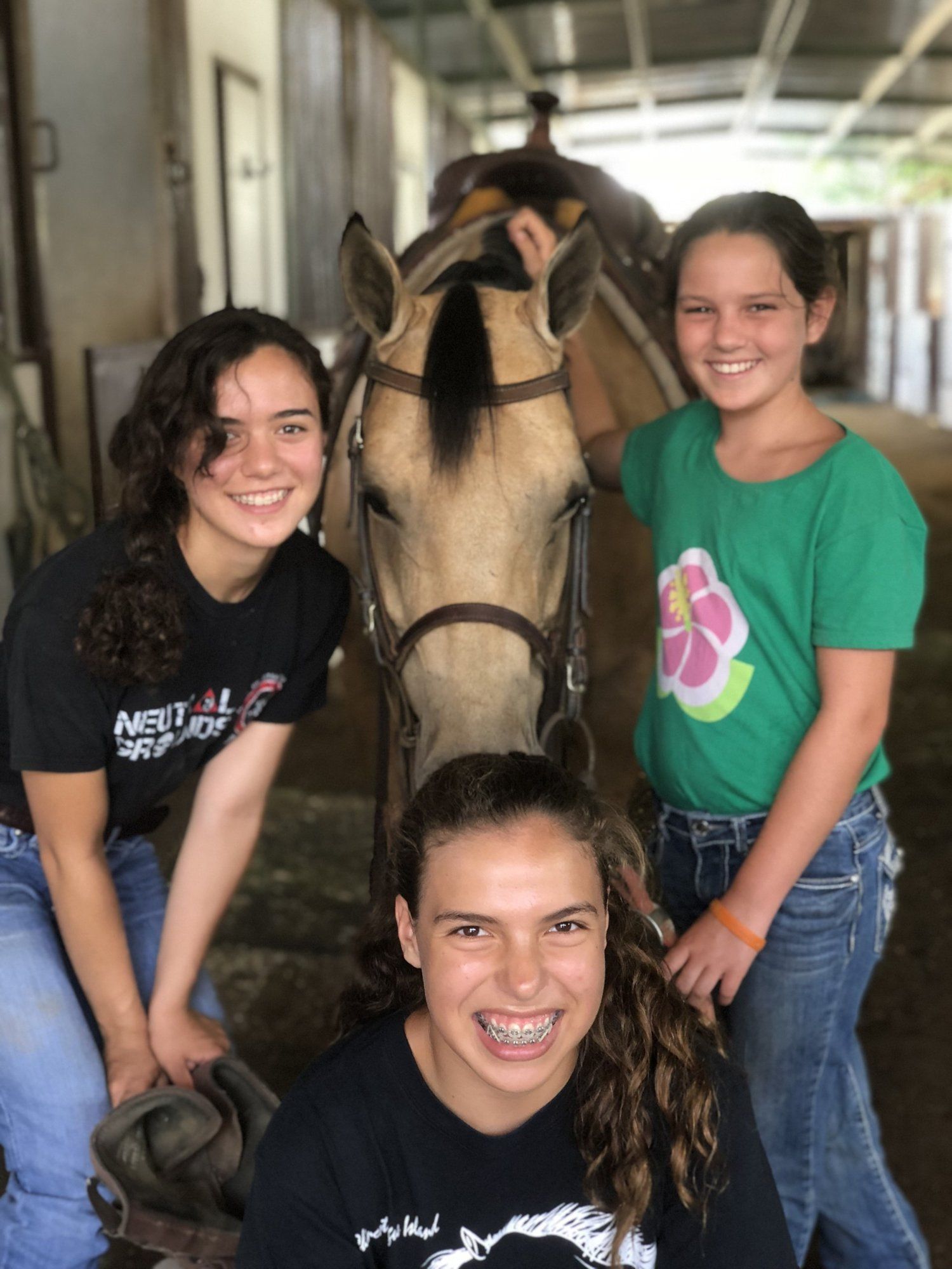 Three girls smiling with a buckskin horse in the barn aisle