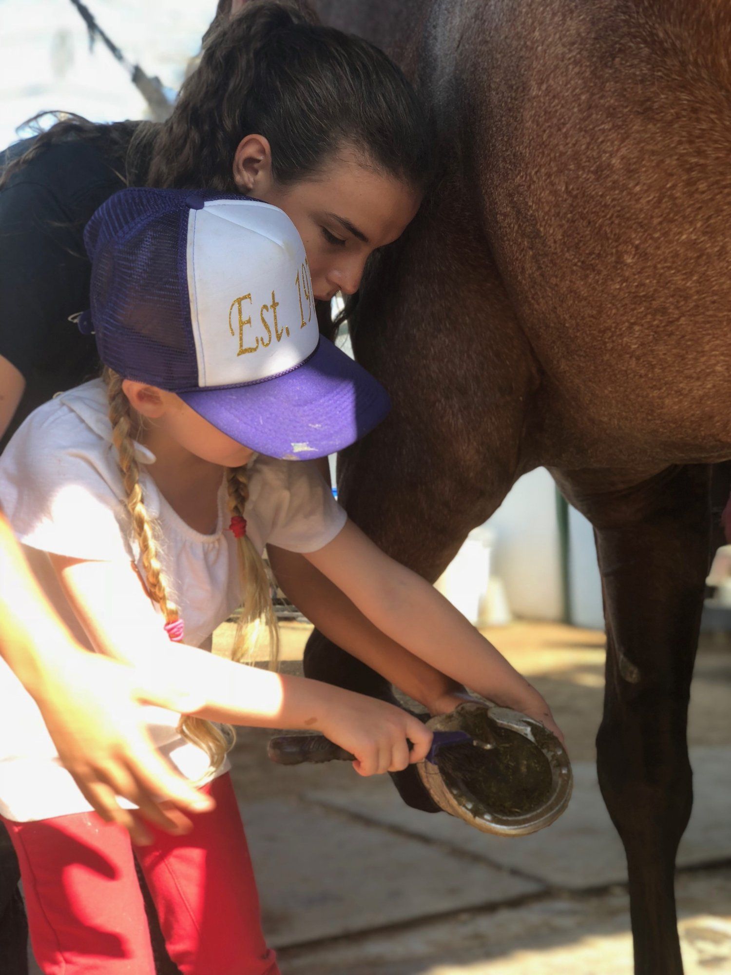 A young girl in a cap working on a horse's hoof while an older girl leans against the horse