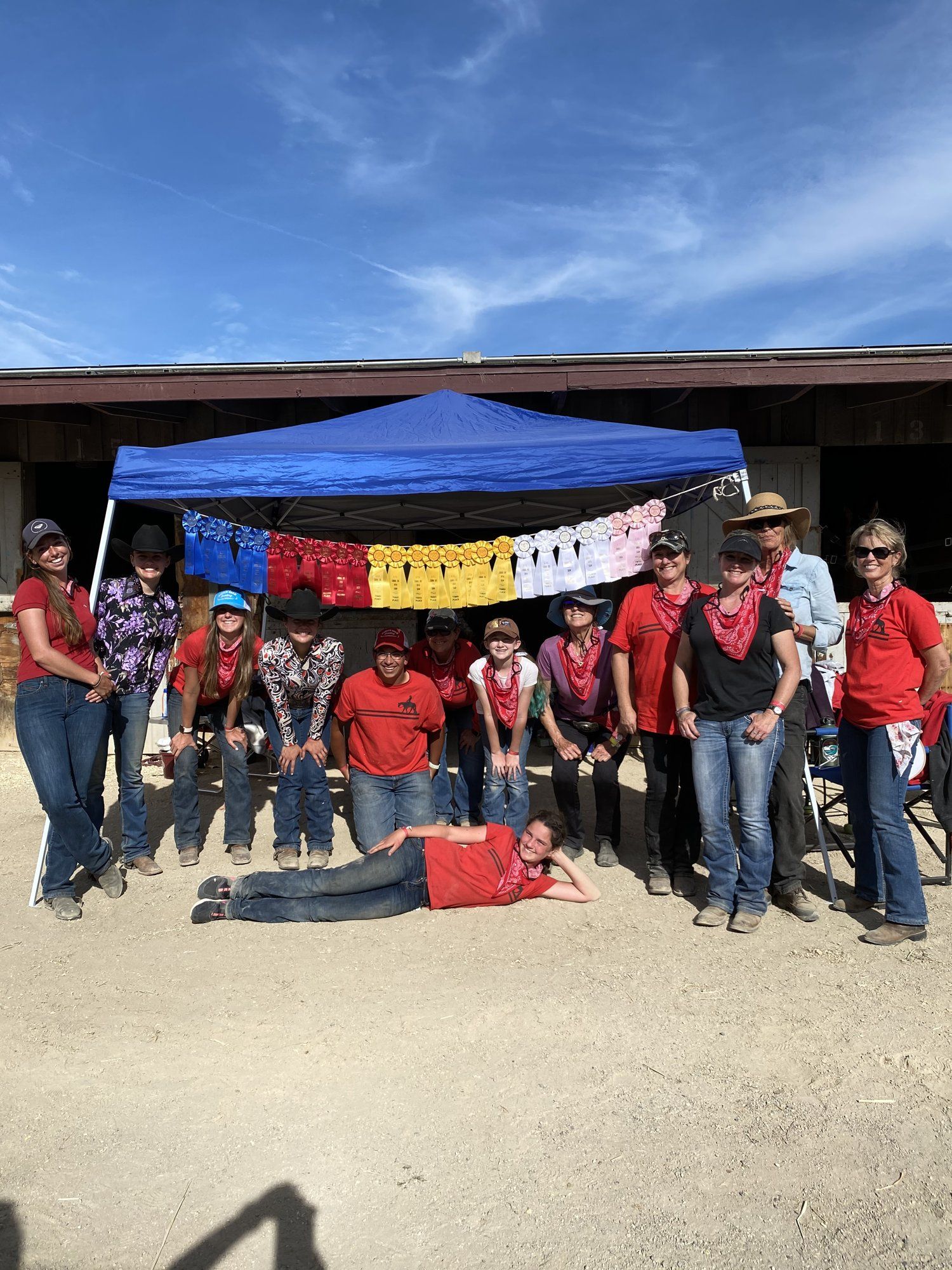 A group in red shirts and bandanas posing under a blue tent at the barn, one person lying on the ground in front