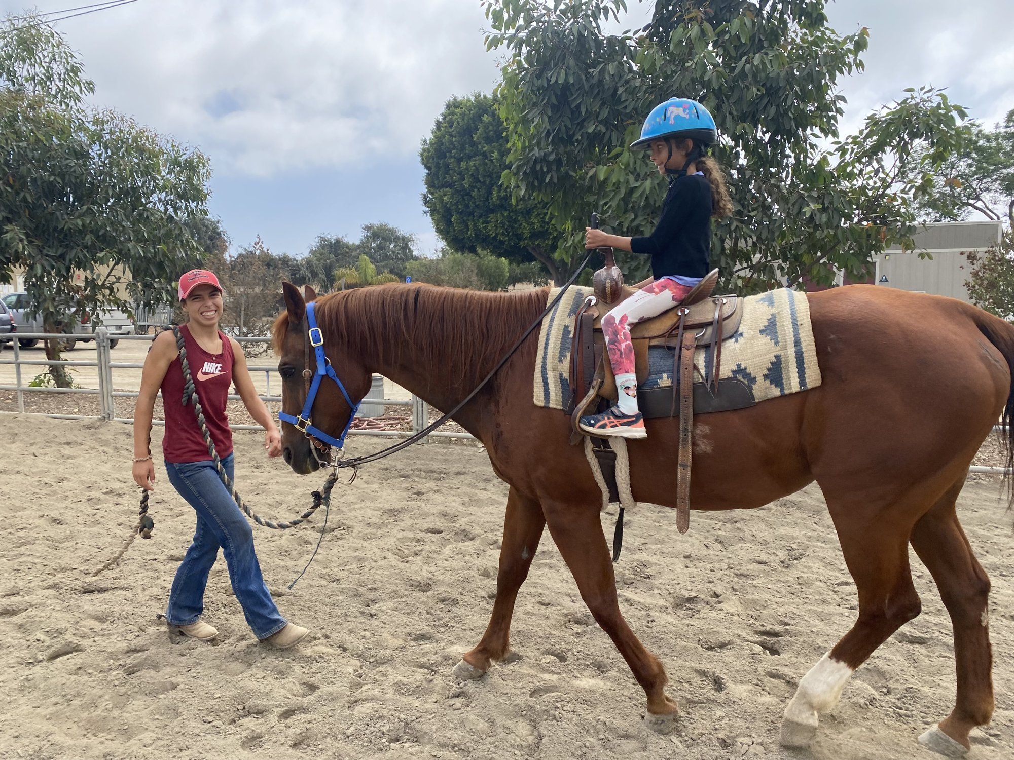 An instructor leads a child in a blue helmet around the arena on a chestnut lesson horse