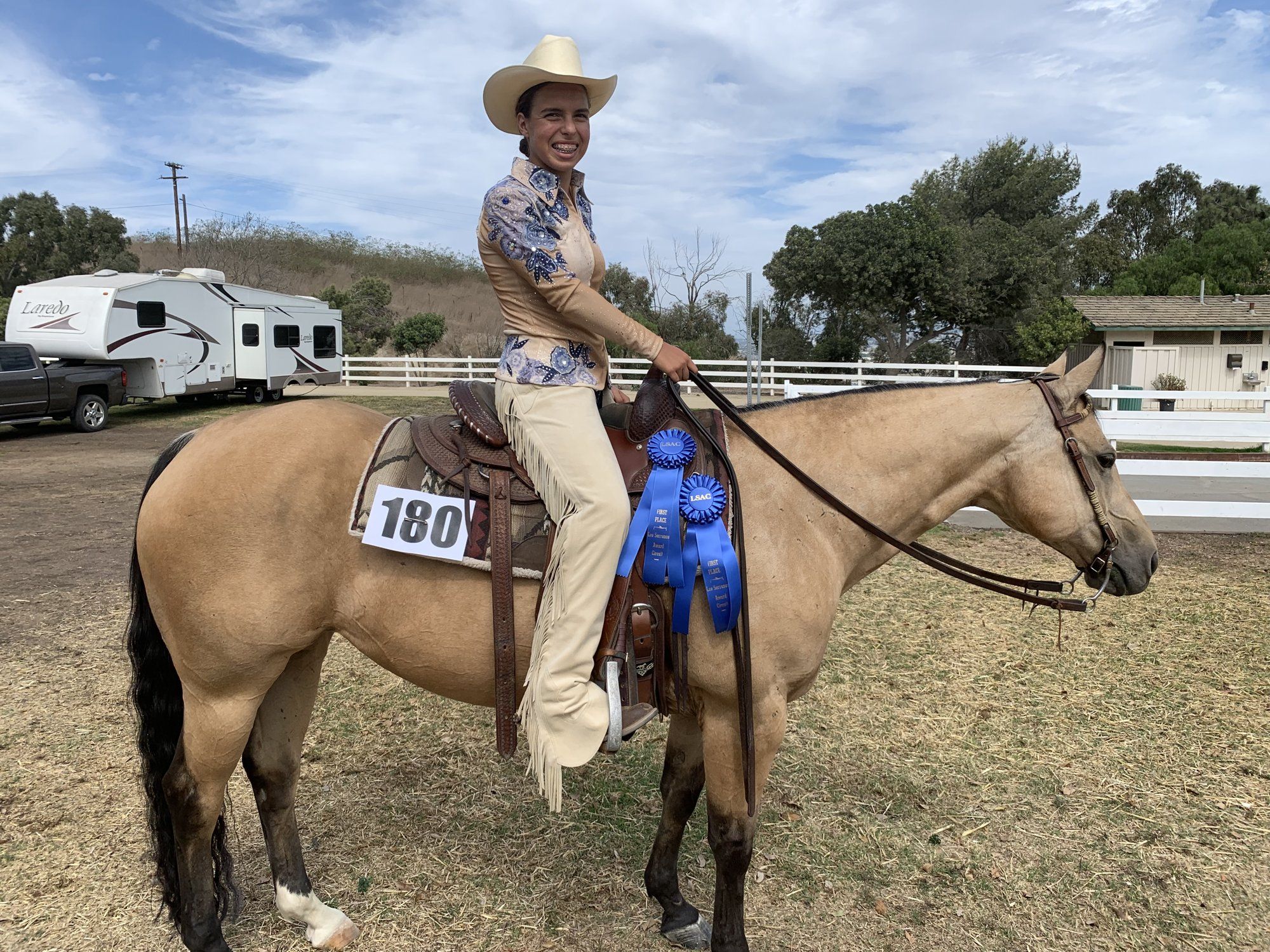 A rider in a cream hat on a buckskin horse at a show, blue ribbons on the saddle